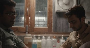 Movie still from “All That Breathes” (2022), directed by Shaunak Sen – Two people are sitting at a table with bottles of alcohol; Medium shot, Over the shoulder angle