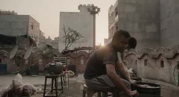 Movie still from “All That Breathes” (2022), directed by Shaunak Sen – A man sitting on top of a stool next to a woman; Wide shot, Low angle