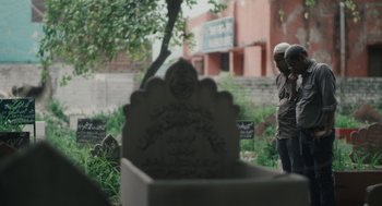 Movie still from “All That Breathes” (2022), directed by Shaunak Sen – Two people standing in front of a grave in a cemetery; Wide shot, Over the shoulder angle