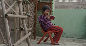 Movie still from “All That Breathes” (2022), directed by Shaunak Sen – A young boy sitting on a chair looking at his phone; Wide shot, High angle
