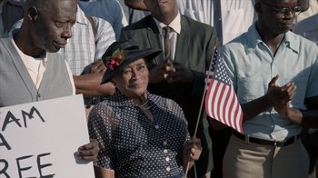 Movie still from “All the Way” (2016), directed by Jay Roach – An older woman holding an american flag in front of a group of people; Medium shot, High angle