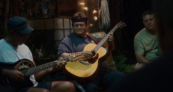 Movie still from “Aloha” (2015), directed by Cameron Crowe – A man holding a guitar while sitting on the ground; Medium shot, Over the shoulder angle