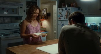 Movie still from “Aloha” (2015), directed by Cameron Crowe – A woman holding an open book in front of a man sitting at a kitchen table; Medium shot, Over the shoulder angle