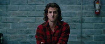 Movie still from “American Animals” (2018), directed by Bart Layton – A man in a red and black plaid shirt sitting in front of a brick wall; Close Up shot, Over the shoulder angle