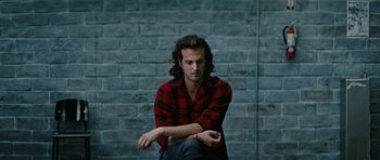Movie still from “American Animals” (2018), directed by Bart Layton – A man sitting on the ground wearing a red and black plaid shirt; Close Up shot, Low angle