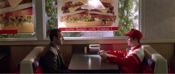 Movie still from “American Beauty” (1999), directed by Sam Mendes – Two people sitting at a table in front of an advertisement; Medium shot, Over the shoulder angle
