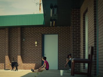 Movie still from “American Honey” (2016), directed by Andrea Arnold – Two people sitting on the ground outside of a brick building; Extreme Wide shot, Over the shoulder angle