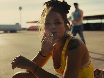 Movie still from “American Honey” (2016), directed by Andrea Arnold – A woman sitting on the ground smoking a cigarette; Medium shot, High angle