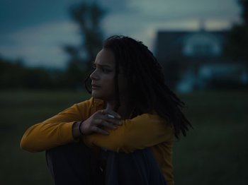 Movie still from “American Honey” (2016), directed by Andrea Arnold – A woman with dreads sitting in a field hugging herself; Medium shot, Low angle
