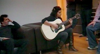Movie still from “Amy” (2015), directed by Asif Kapadia – A woman sitting on a couch playing an acoustic guitar; Wide shot, High angle