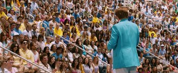 Movie still from “Anchorman 2: The Legend Continues” (2013), directed by Adam McKay – A crowd of people sitting on bleachers watching a man speak; Wide shot, High angle