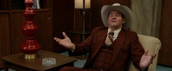 Movie still from “Anchorman 2: The Legend Continues” (2013), directed by Adam McKay – A man in a suit and a cowboy hat is sitting at a table; Medium shot, Over the shoulder angle