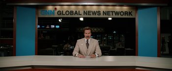 Movie still from “Anchorman 2: The Legend Continues” (2013), directed by Adam McKay – Anchorman ron burgundy sitting at a news desk in front of a sign; Medium shot, Low angle