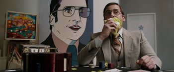 Movie still from “Anchorman 2: The Legend Continues” (2013), directed by Adam McKay – A man sitting in front of a painting eating a sandwich; Medium shot, Over the shoulder angle