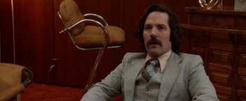 Movie still from “Anchorman 2: The Legend Continues” (2013), directed by Adam McKay – A man in a suit sitting in front of a chair; Close Up shot, Over the shoulder angle