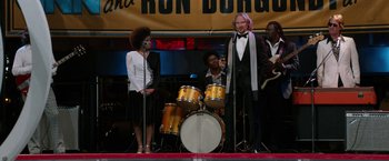 Movie still from “Anchorman 2: The Legend Continues” (2013), directed by Adam McKay – A group of people standing on a stage next to drums; Medium shot, Over the shoulder angle