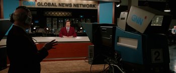 Movie still from “Anchorman 2: The Legend Continues” (2013), directed by Adam McKay – Anchorman impersonator dressed as ron burgundy sitting at a news desk; Wide shot, Over the shoulder angle