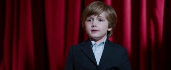 Movie still from “Anchorman 2: The Legend Continues” (2013), directed by Adam McKay – A young boy in a suit stands in front of a red curtain; Close Up shot, High angle