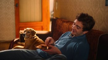 Movie still from “And Then We Danced” (2019), directed by Levan Akin – A man sitting on a couch with a plate of food in front of him and a dog; Medium shot, Over the shoulder angle