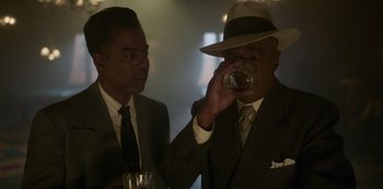 Movie still from “Fargo” (2014), created by Noah Hawley – A man in a suit and a hat is drinking a glass of wine; Medium shot, Over the shoulder angle