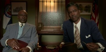 Movie still from “Fargo” (2014), created by Noah Hawley – Two men in suits sitting at a table in front of a stained glass window; Medium shot, Over the shoulder angle