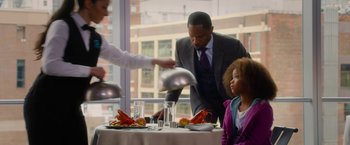 Movie still from “Annie” (2014), directed by Will Gluck – A man in a suit serves a little girl at a table; Medium shot, Over the shoulder angle