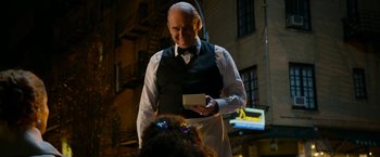 Movie still from “Annie” (2014), directed by Will Gluck – An older man in a vest and bow tie holding a card; Medium shot, Over the shoulder angle