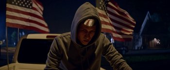 Movie still from “Assassination Nation” (2018), directed by Sam Levinson – A man in a hooded sweatshirt with an american flag in the background; Medium shot, Low angle