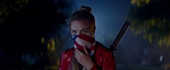 Movie still from “Assassination Nation” (2018), directed by Sam Levinson – A woman in a red jacket holding a baseball bat in front of her face with an american flag mask on; Close Up shot, Over the shoulder angle