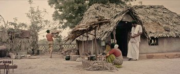Two women sit outside of a thatched roof hut