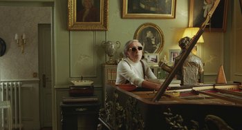 Movie still from “Attila Marcel” (2013), directed by Sylvain Chomet – An older woman sitting at a desk in front of a painting on the wall; Medium shot, Over the shoulder angle