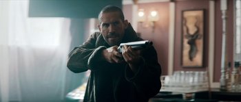 Movie still from “Avengement” (2019), directed by Jesse V. Johnson – A man holding a gun while wearing a brown coat; Medium shot, Over the shoulder angle