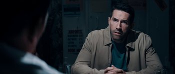 Movie still from “Avengement” (2019), directed by Jesse V. Johnson – A man sitting at a table in front of an airport sign; Close Up shot, Over the shoulder angle