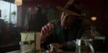 Movie still from “Fargo” (2014), created by Noah Hawley – An older man sitting at a table with a cup of coffee; Medium shot, Low angle