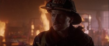 Movie still from “Backdraft” (1991), directed by Ron Howard – A man in a fireman's uniform is looking at the camera while standing in front of a burning building; Close Up shot, Low angle