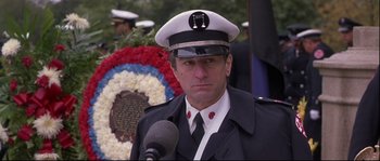 Movie still from “Backdraft” (1991), directed by Ron Howard – A man in a uniform is speaking into a microphone in front of a wreath; Close Up shot, Low angle