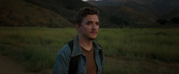 Movie still from “Band of Robbers” (2015), directed by Adam Nee – A young man standing in front of a grassy field; Close Up shot, Over the shoulder angle
