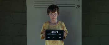 Movie still from “Band of Robbers” (2015), directed by Adam Nee – A young boy holding up a mug shot in front of a wall; Medium shot, Low angle