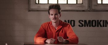 Movie still from “Band of Robbers” (2015), directed by Adam Nee – A man in an orange shirt sitting at a table in a jail cell; Close Up shot, High angle
