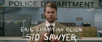 Movie still from “Band of Robbers” (2015), directed by Adam Nee – A man in a suit and tie with a sign in front of a fence; Medium shot, Over the shoulder angle