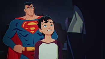 Movie still from “Batman and Superman: Battle of the Super Sons” (2022), directed by Matt Peters – A boy standing next to a man wearing a superman costume; Medium shot, Over the shoulder angle