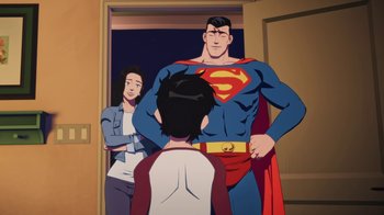 Movie still from “Batman and Superman: Battle of the Super Sons” (2022), directed by Matt Peters – A man in a superman costume standing in front of a woman and a boy; Medium shot, Over the shoulder angle