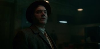 Movie still from “Fargo” (2014), created by Noah Hawley – A man wearing a suit and a hat is standing in a dark room; Medium shot, Low angle