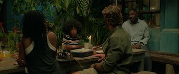 Movie still from “Beast” (2022), directed by Baltasar Kormákur – A group of people sitting at a table with food; Medium shot, Over the shoulder angle