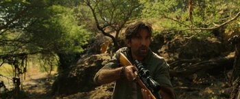 Movie still from “Beast” (2022), directed by Baltasar Kormákur – A man holding a rifle in a wooded area; Medium shot, Over the shoulder angle