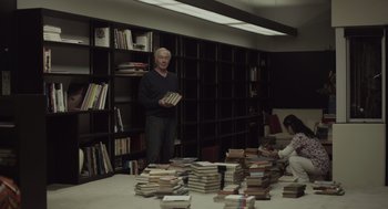 Movie still from “Beginners” (2010), directed by Mike Mills – A man standing in front of a pile of books in a room; Medium shot, High angle