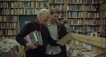 Movie still from “Beginners” (2010), directed by Mike Mills – Two men standing next to each other in front of shelves of books; Medium shot, Over the shoulder angle