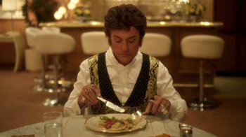 Movie still from “Behind the Candelabra” (2013), directed by Steven Soderbergh – A person sitting at a table with a plate of food; Medium shot, Over the shoulder angle