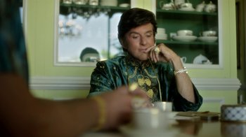 Movie still from “Behind the Candelabra” (2013), directed by Steven Soderbergh – A woman sitting at a table with a cup of coffee; Medium shot, Over the shoulder angle
