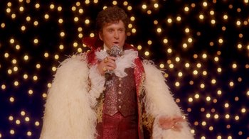 Movie still from “Behind the Candelabra” (2013), directed by Steven Soderbergh – A man in a red and white outfit holding a microphone; Medium shot, Over the shoulder angle